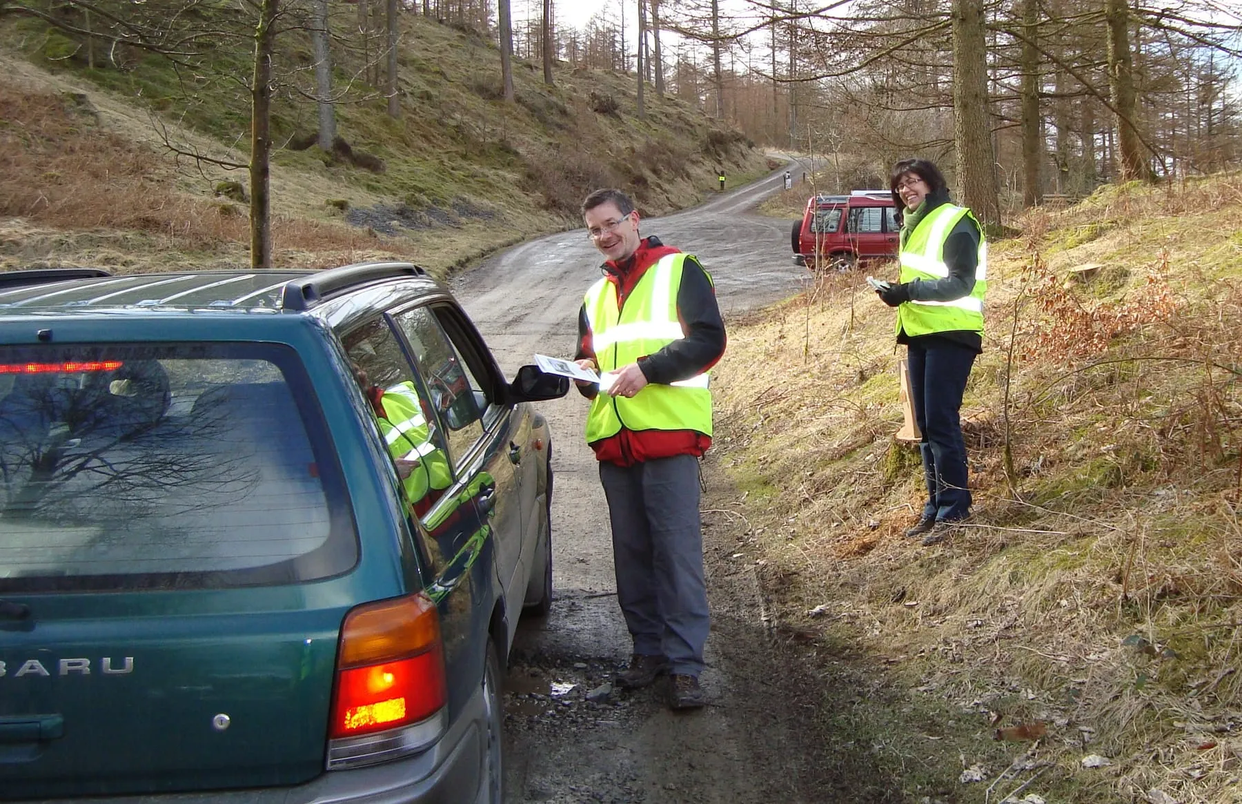 Two people in high-visibility vests directing a car on a forest track