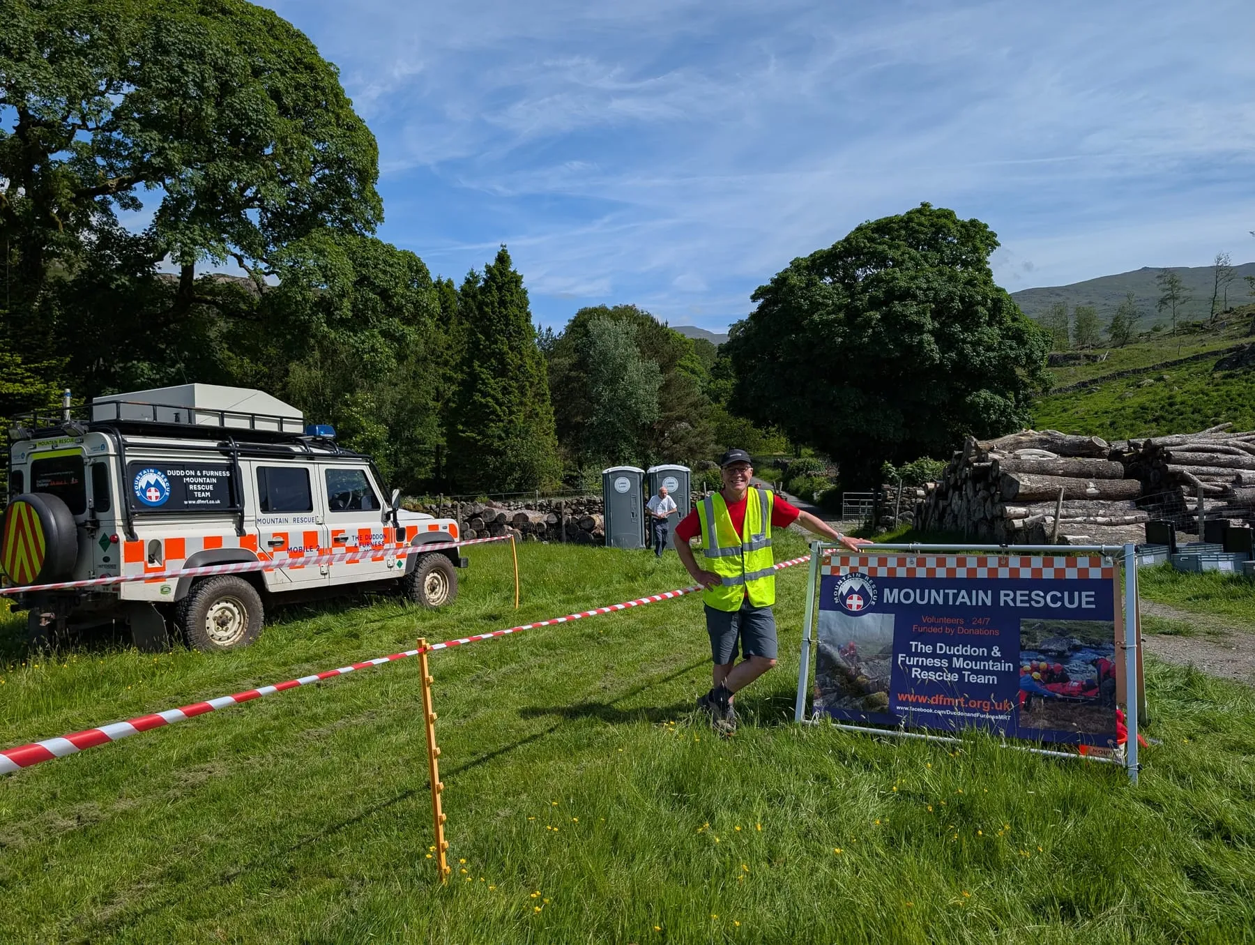 A team member in a high-visibility vest beside a Land Rover and a Mountain Rescue banner at an outdoor event