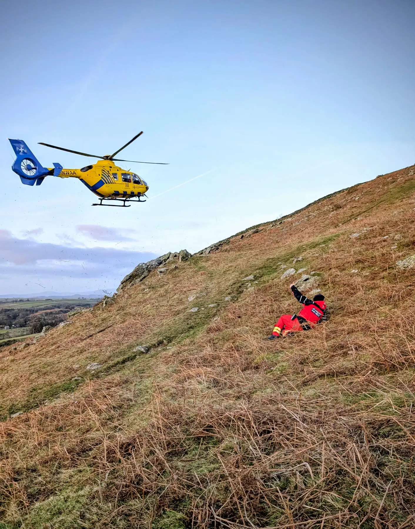A helicopter hovering above a team member on a bracken-covered fellside