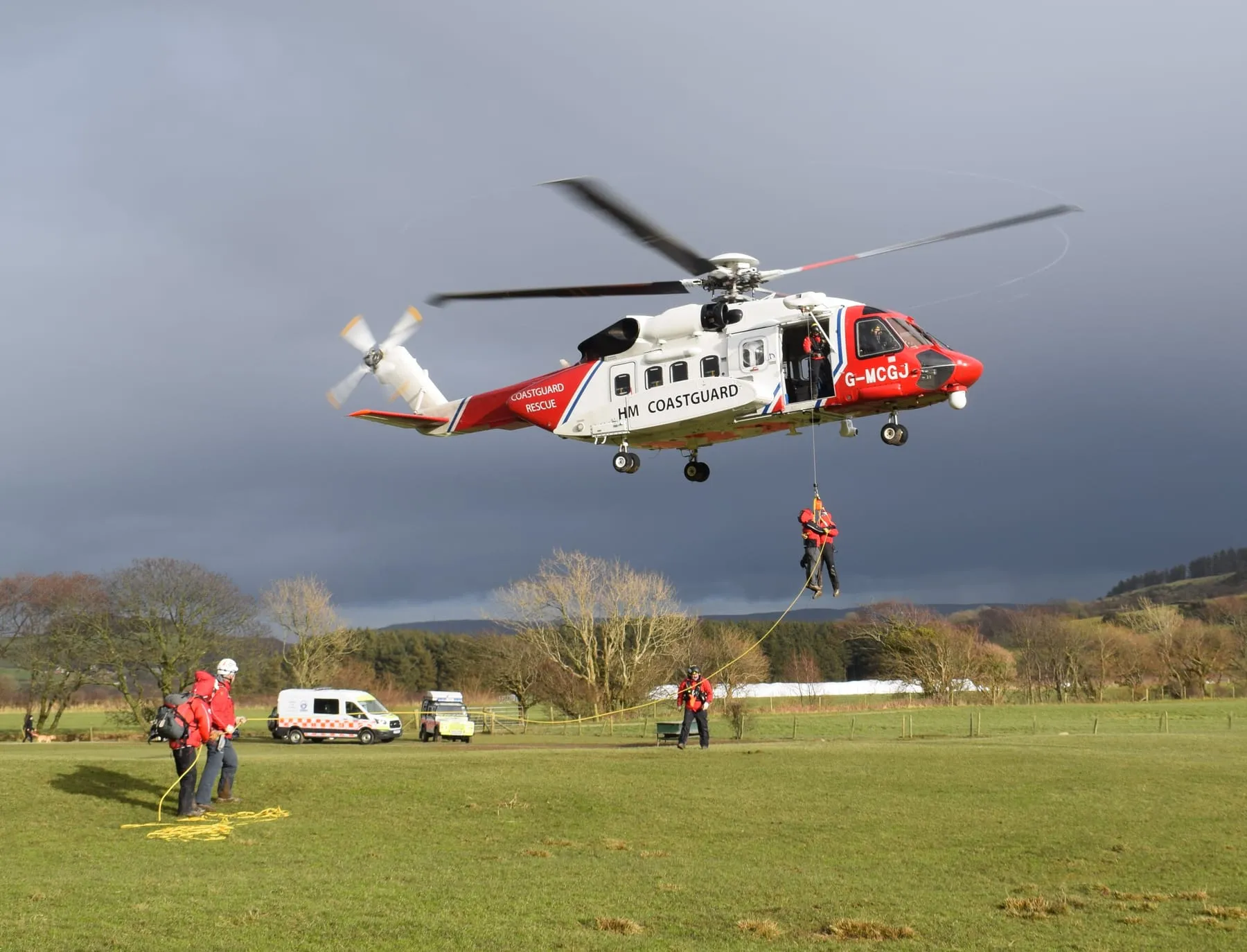 A person being winched from an HM Coastguard helicopter above a field while team members hold a line