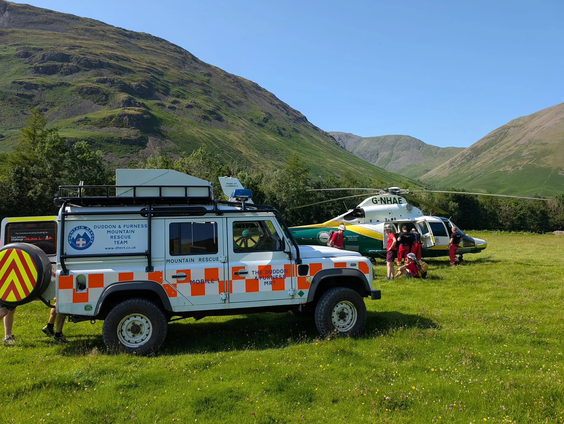 A team Land Rover beside a helicopter and its crew in a valley