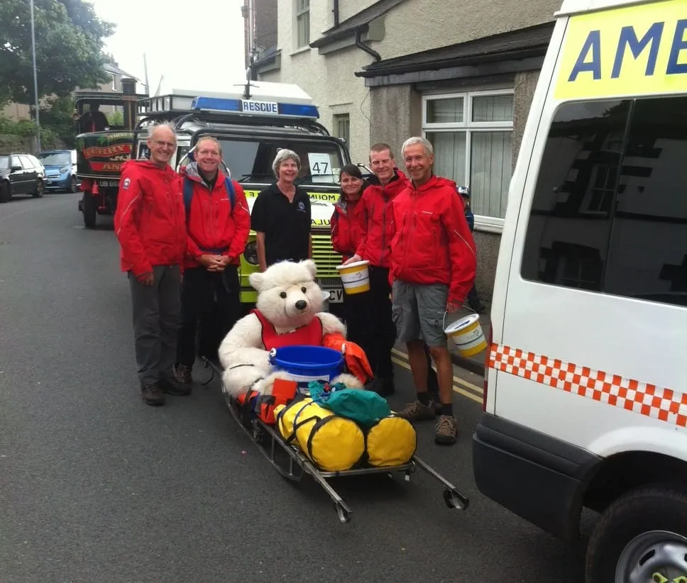 Team members with collecting buckets and a teddy bear on a stretcher beside a team ambulance