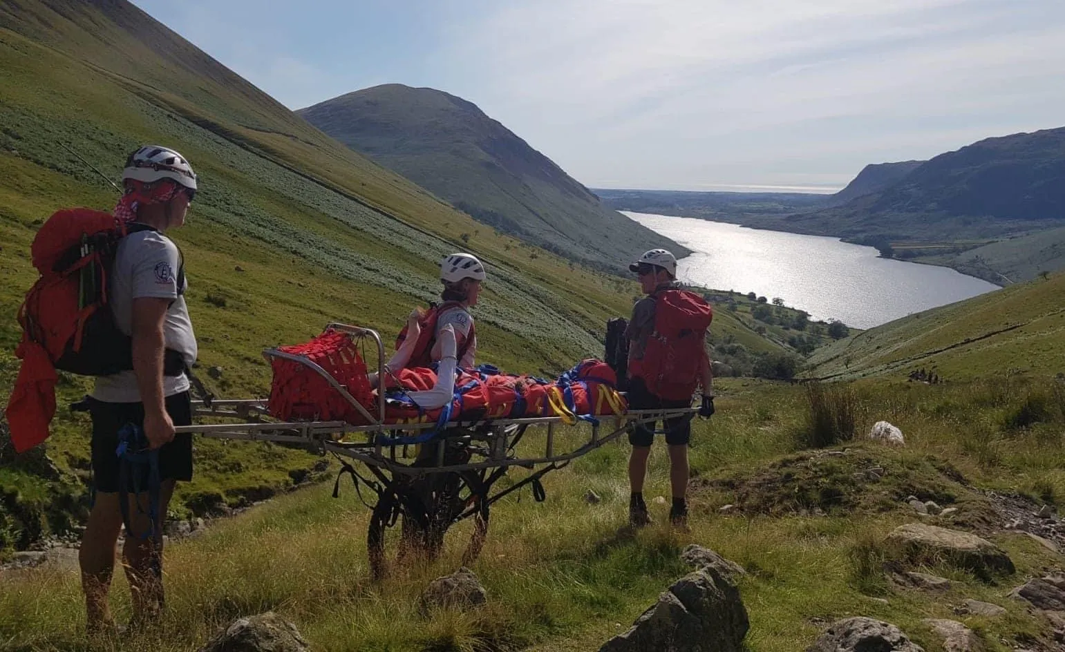 Team members wheeling a stretcher down a valley path above a lake